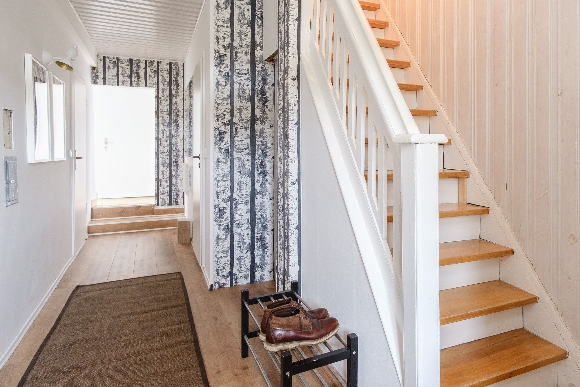 Una escalera con peldaños de madera y barandillas blancas, que conduce a un rellano con una puerta y un muro con papel tapiz de diseño vertical.