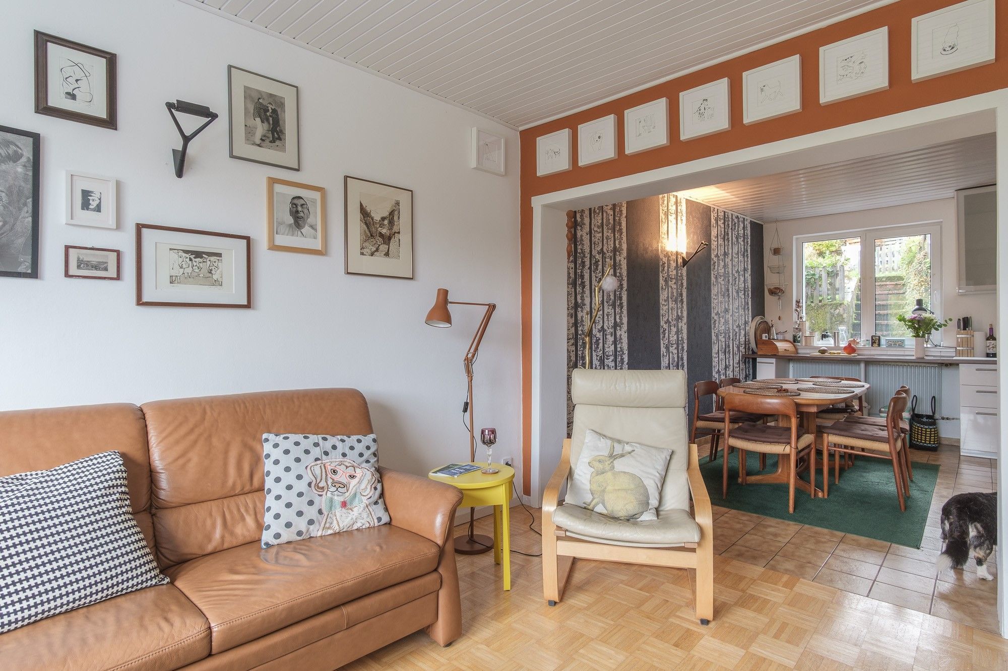 Uma sala de estar com um sofá de couro marrom, uma mesinha lateral amarela e uma cadeira com almofada de coelho. Há quadros nas paredes e uma janela com vista para o exterior.