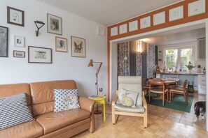 A living room with a brown leather sofa, a yellow side table, and a chair with a rabbit pillow. There are framed pictures on the wall and a window with a view of the outside. - Haushimmelblau, Germany (Sankt Andreasberg)