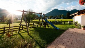 A playground with a slide and swings, surrounded by a wooden fence, grassy area, and a brick pathway. - Apartment 16 - Liedschreiber GbR Guesthouse and Vacation Rentals (Kreuth)