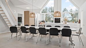 Un comedor moderno con una mesa grande, sillas negras e iluminación colgante. Se ve una escalera a la izquierda y una zona de cocina al fondo.