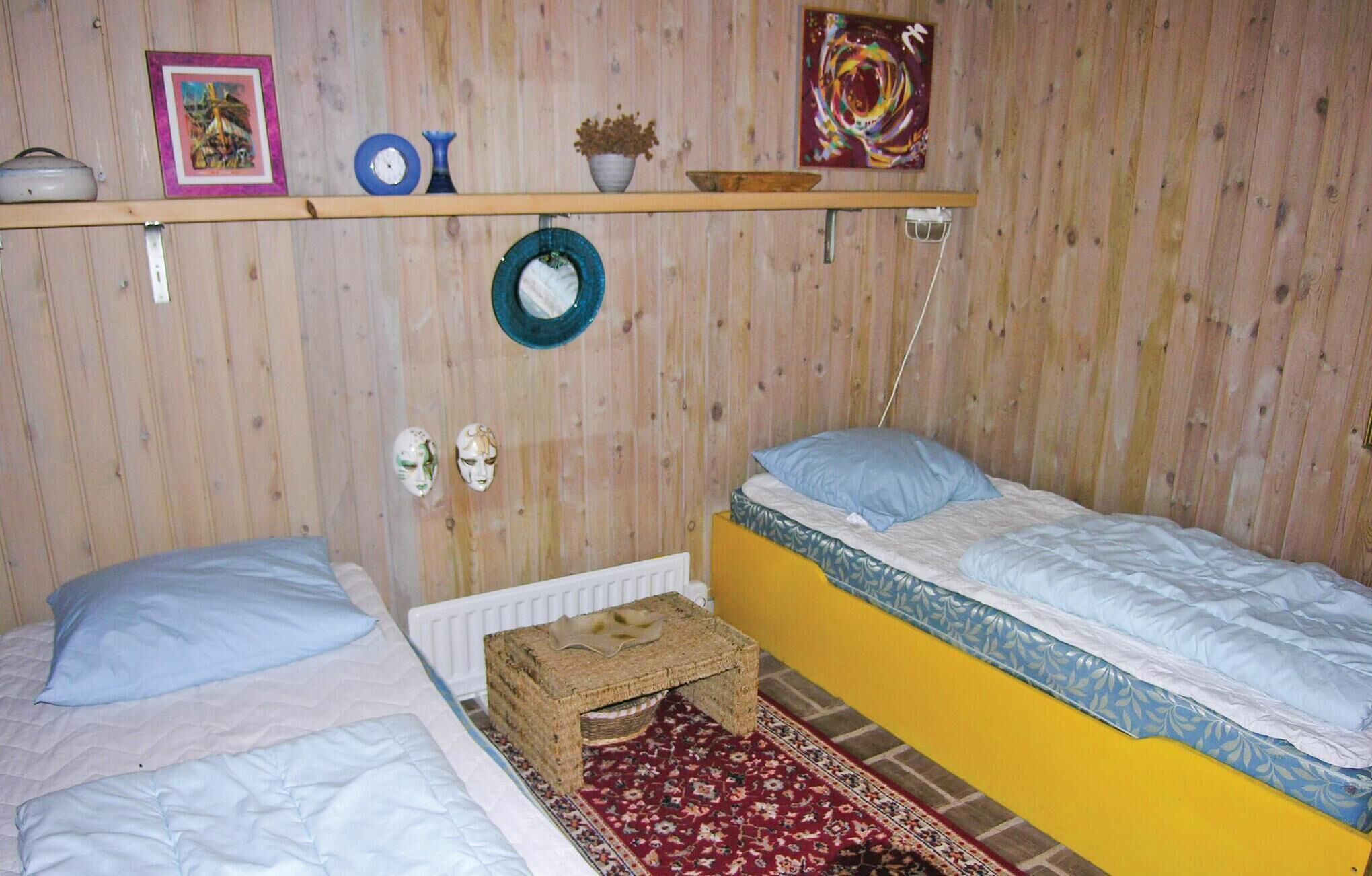A room with two beds, a wooden wall, a shelf with various items, and a red patterned rug.