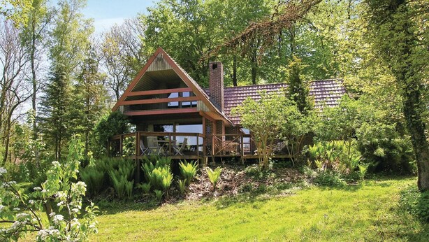 Un chalet rustique doté d’une terrasse en bois et d’une cheminée en briques, entouré d’une végétation luxuriante.
