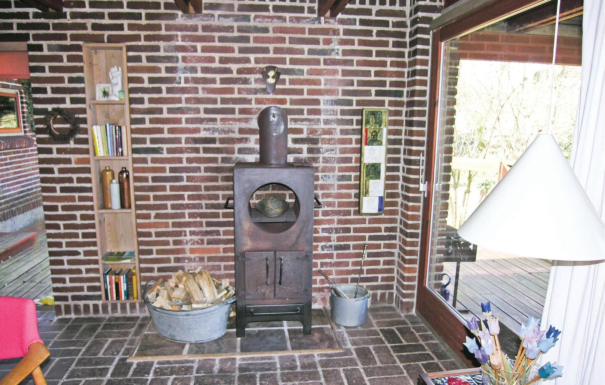 Een rustieke kamer met een met baksteen beklede open haard, een houten plank met boeken, een metalen emmer met houtblokken en een raam met uitzicht op bomen.