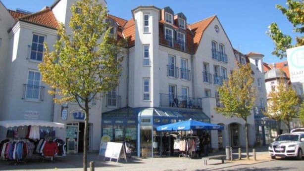 A row of white buildings with red-tiled roofs, a storefront with a blue awning, and a sign that reads "Stadtbücherei." - (146) 2-room apartment - Apartment house Olymp (Kühlungsborn)