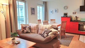 A modern living room with a brown sofa, wooden coffee table, and a dining area with a red kitchen in the background. - (146) 2-room apartment - Apartment house Olymp (Kühlungsborn)