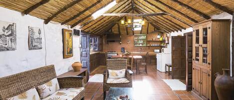 A rustic interior with a high thatched roof, wicker furniture, a glass coffee table, and a kitchen area with wooden cabinets.