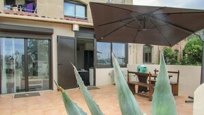 Een patio met een tafel, stoelen, een grote parasol en hoge agaveplanten.
