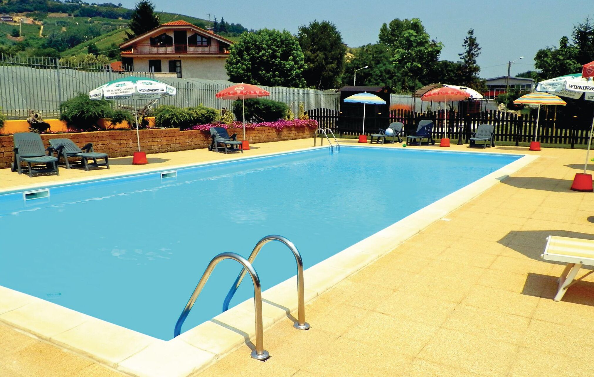 A swimming pool with lounge chairs and umbrellas, surrounded by a fence and a building in the background.