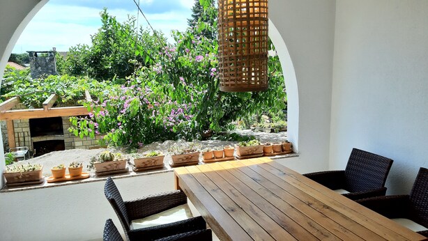 Une terrasse extérieure avec une table en bois et des chaises en osier, entourée de plantes en pot et agrémentée d’une cheminée en pierre.