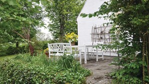 A white building with a garden area featuring a white bench and table, surrounded by greenery and a small flower bed.