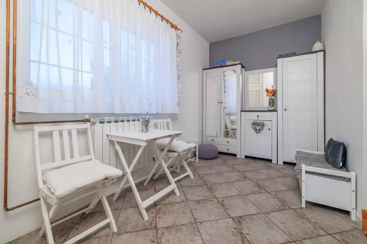 A room with a white bed, a small table, a chair, a cabinet, and a window with curtains.