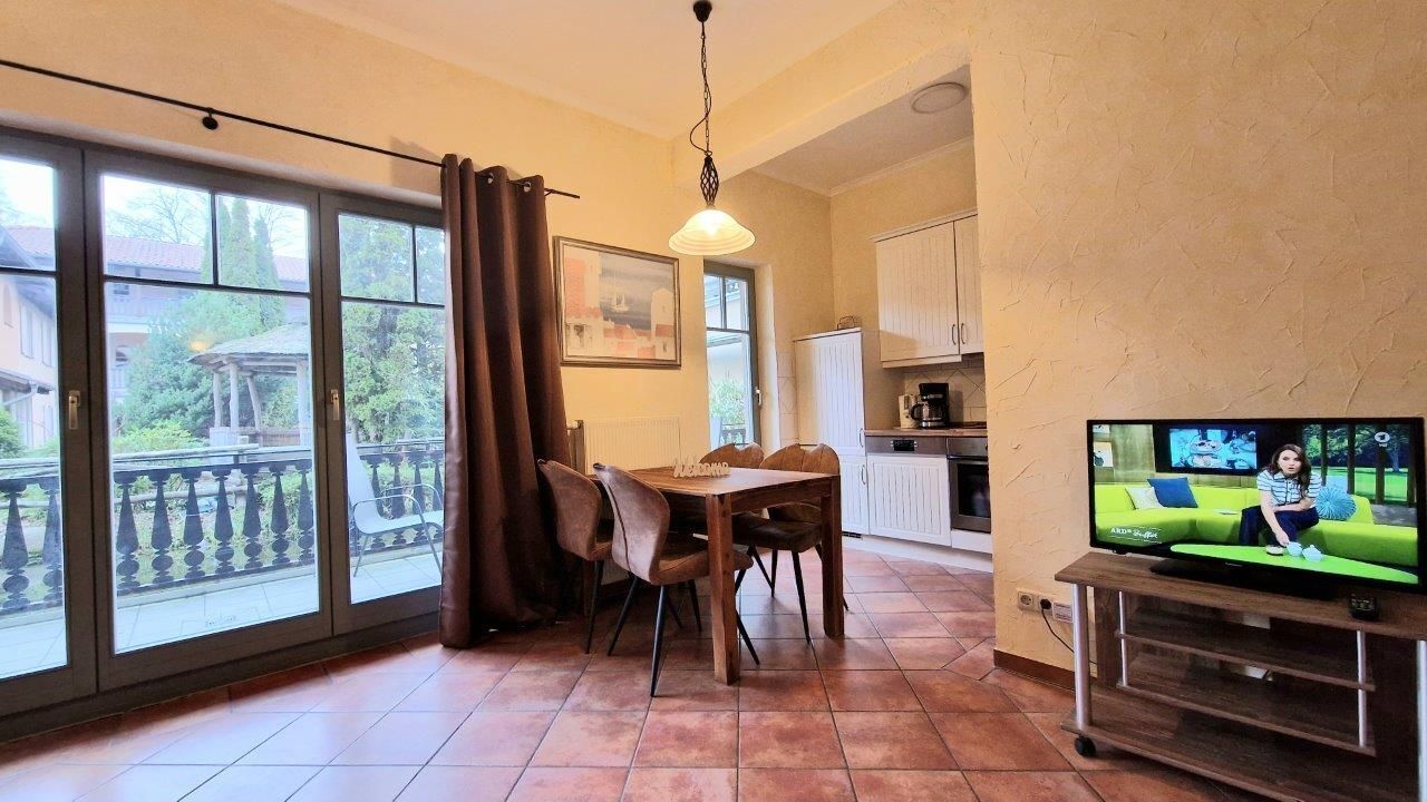 Un comedor con una mesa y sillas, un televisor sobre un mueble bajo, y vistas a un balcón con barandilla.