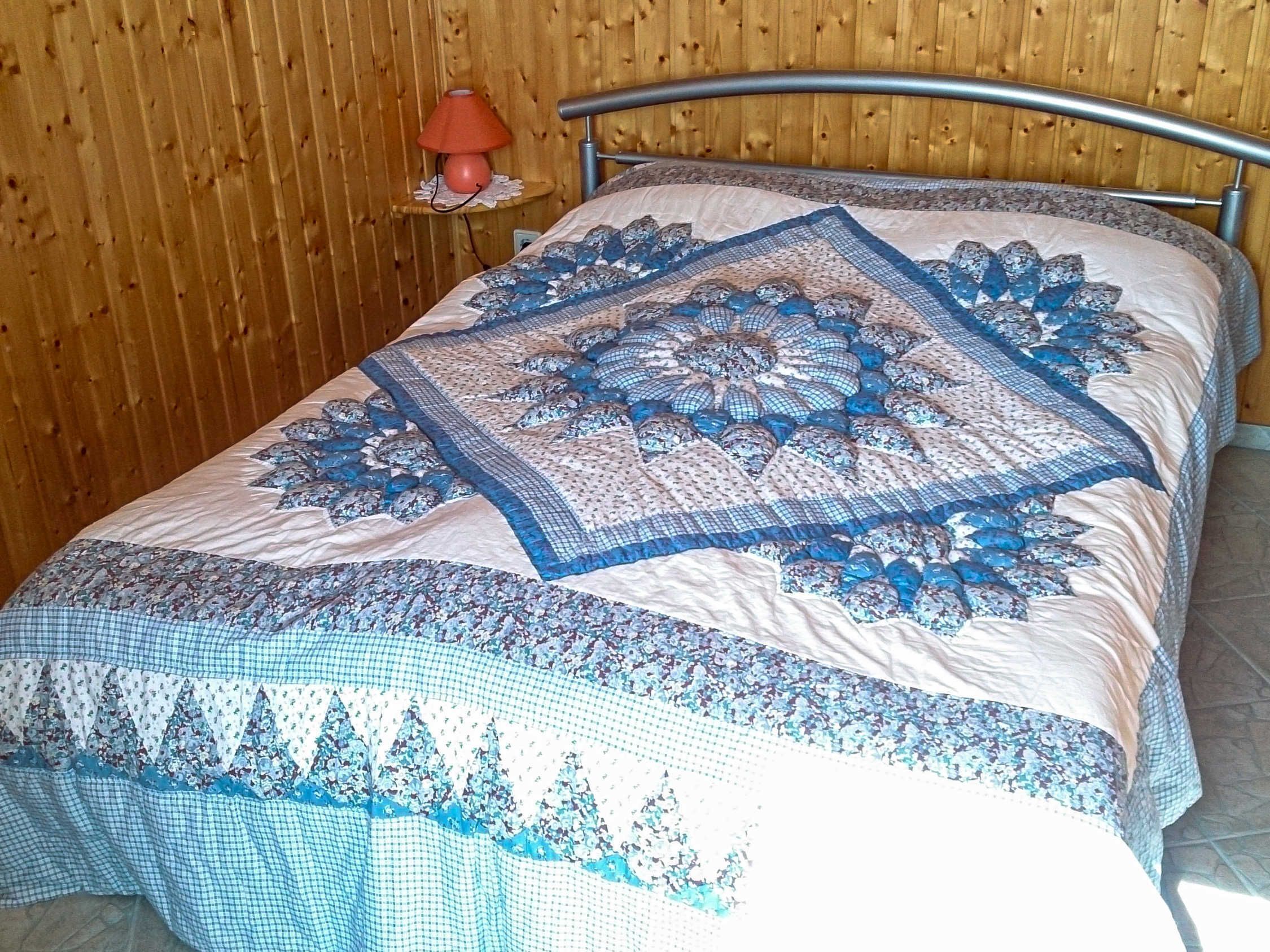 Ein Bett mit blau-weißer Bettdecke und Kissen, eine Holzwand und ein kleiner Tisch mit einer Lampe.