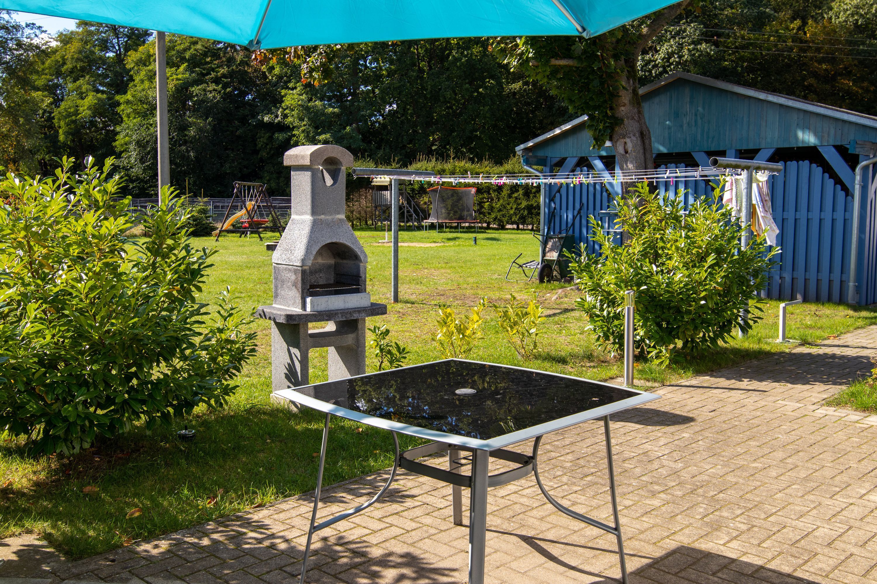 A patio area with a glass-top table, a stone barbecue grill, and a blue shed in the background.