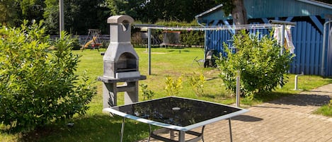 A patio area with a glass-top table, a stone barbecue grill, and a blue shed in the background.