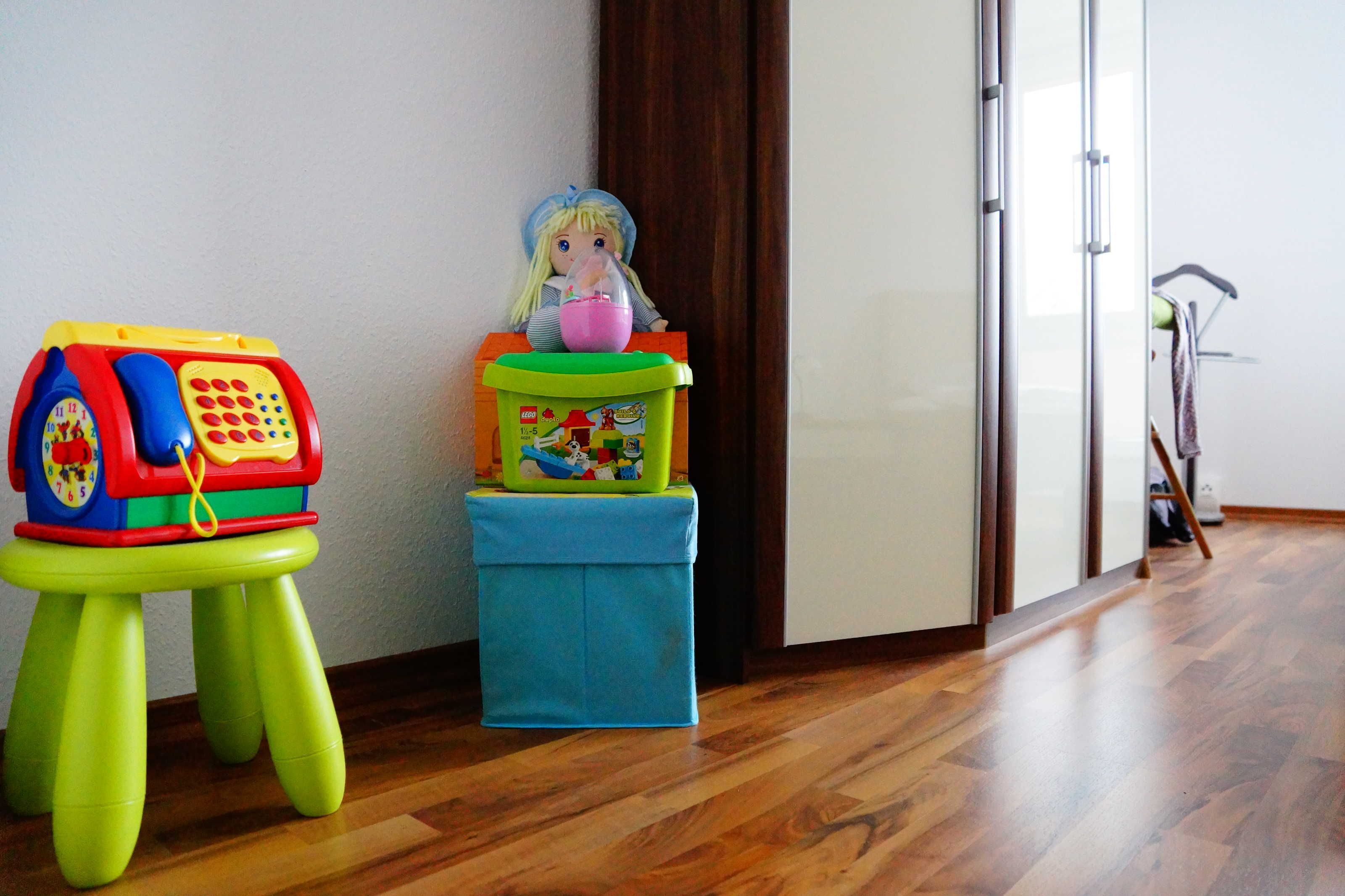 A doll on a stack of colorful toy stools in a room with wooden flooring and a white door in the background.