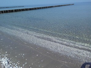 En strand med klart vatten, småsten och en träbrygga.