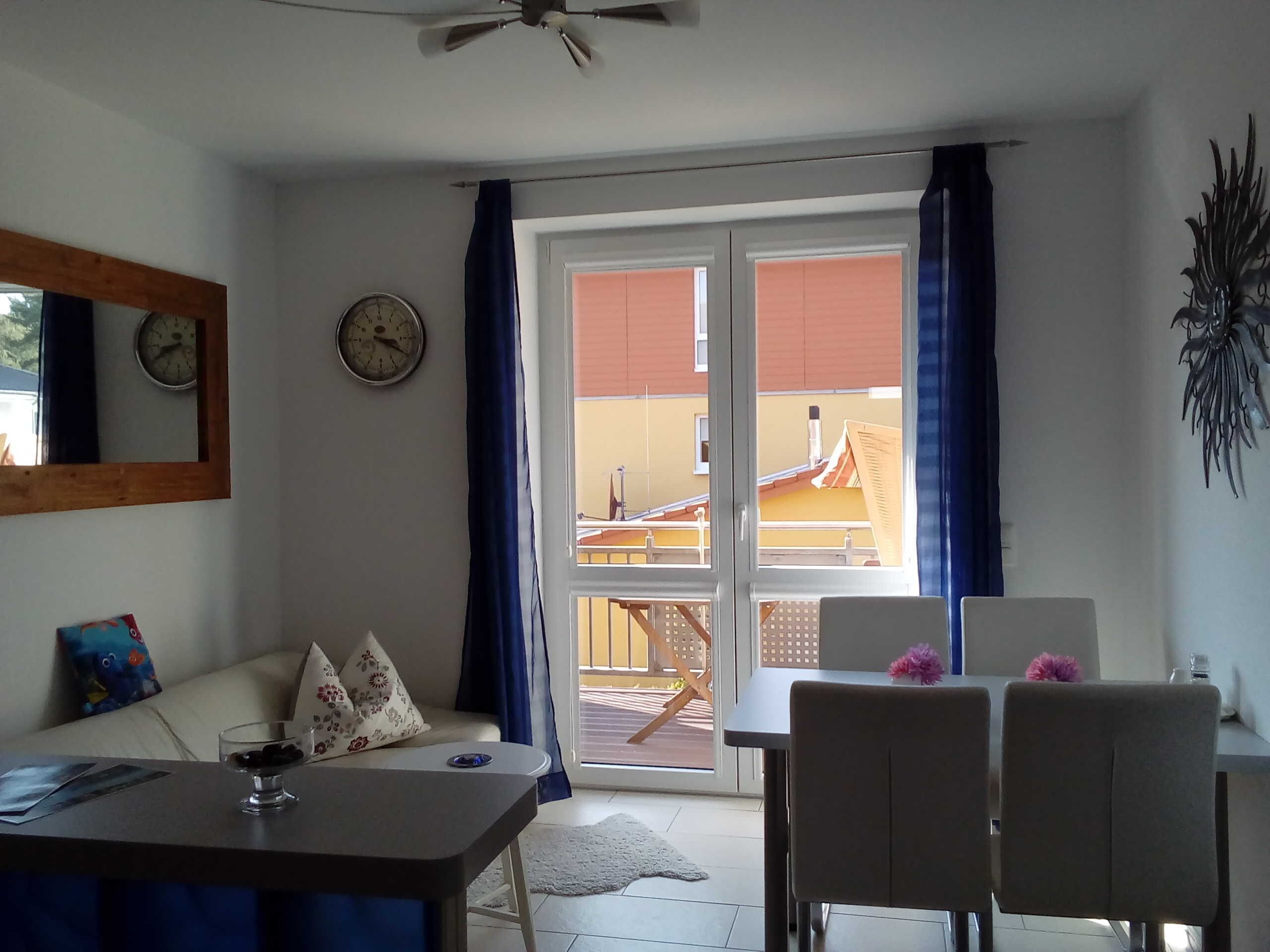 Een kamer met een eettafel, stoelen, een bank, een klok en uitzicht door een glazen deur op een buitenbalkon.