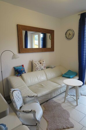 Una sala de estar con un sofá de cuero blanco, una mesa redonda, un sillón blanco, un espejo, un reloj y una cortina azul.