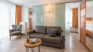 Una habitación de hotel moderna con sofá, sillón y una pequeña mesa de madera con un florero y una revista.