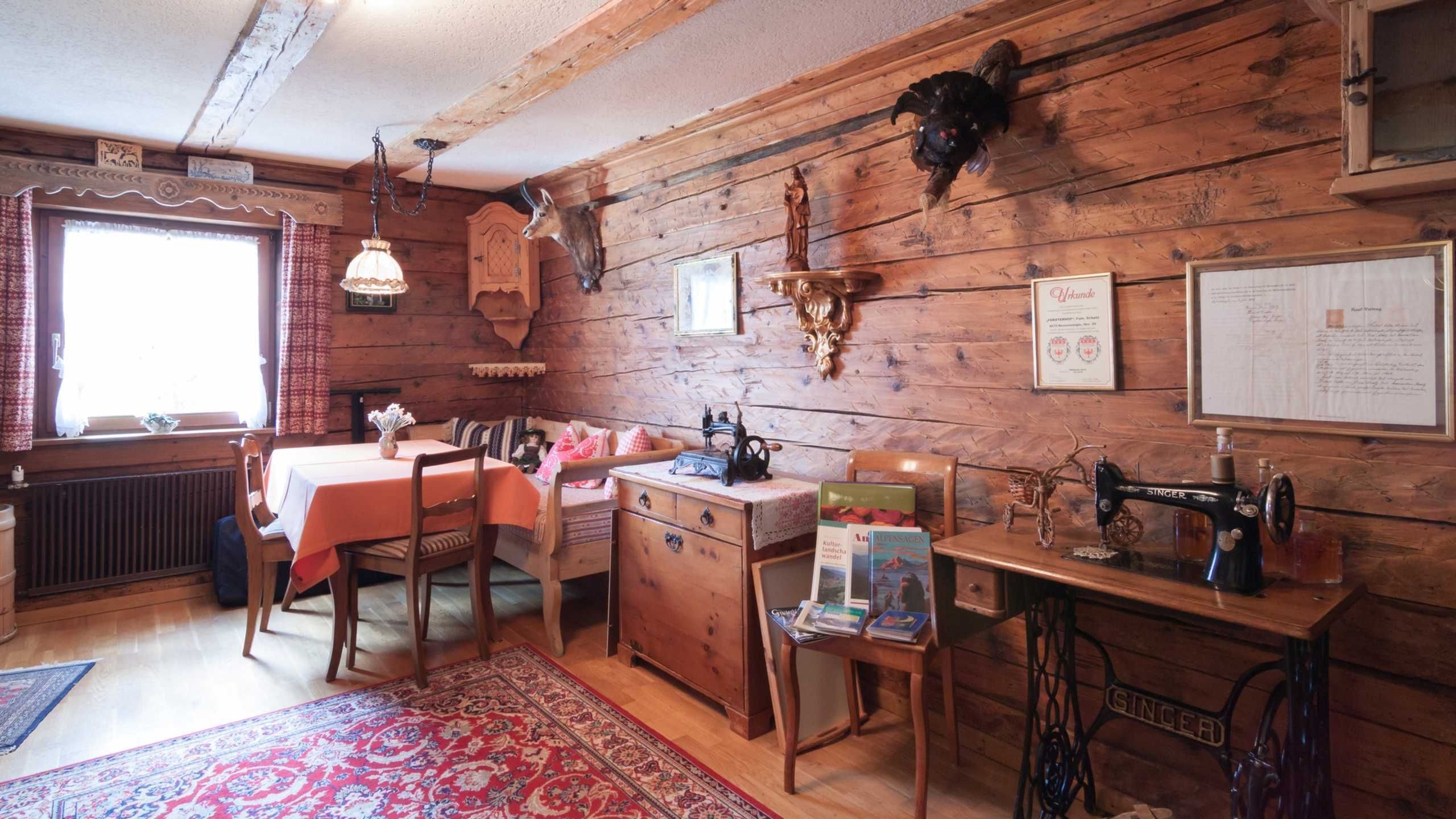 A rustic room with wooden walls, a dining table, chairs, a radiator, a sewing machine, and a window with curtains.