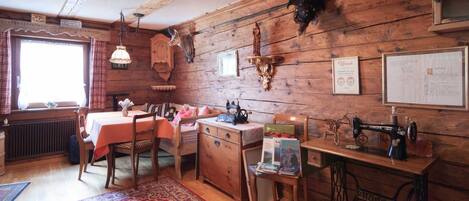 A rustic room with wooden walls, a dining table, chairs, a radiator, a sewing machine, and a window with curtains.