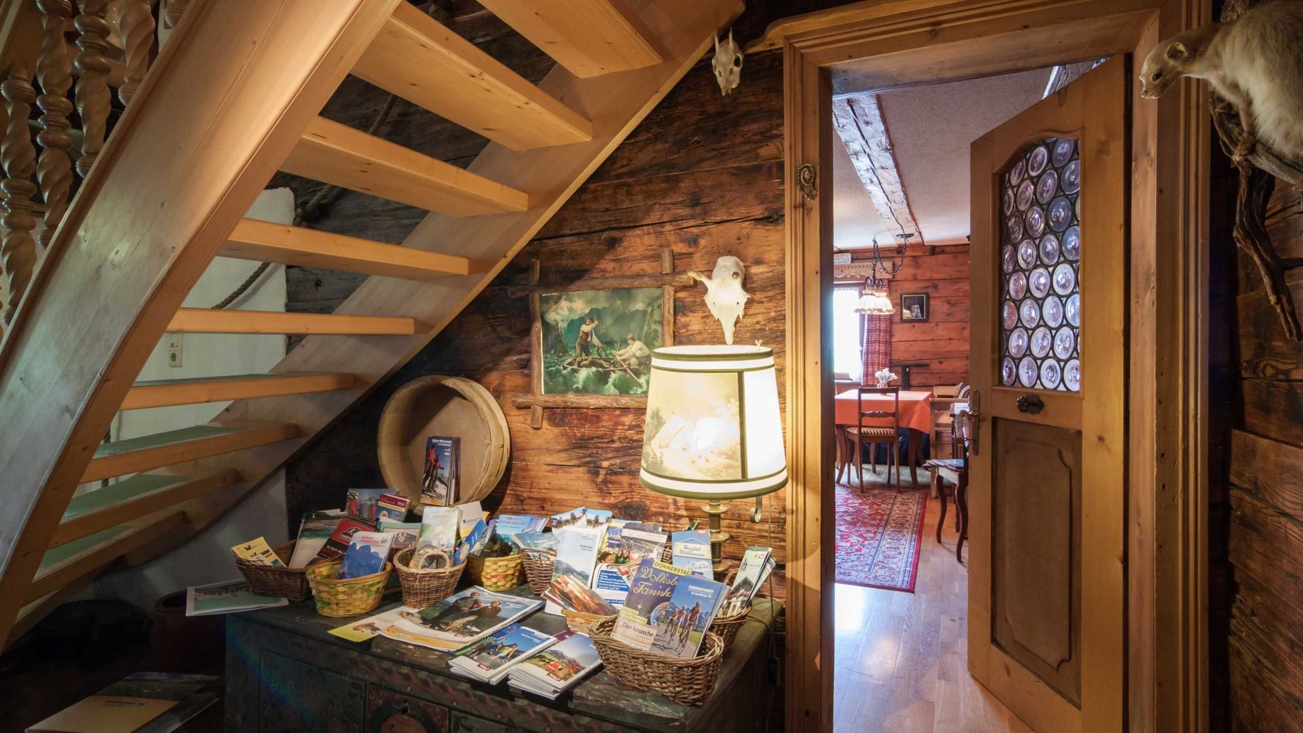 A rustic room with a wooden staircase, a table with postcards, a lamp, and a door leading to another room.