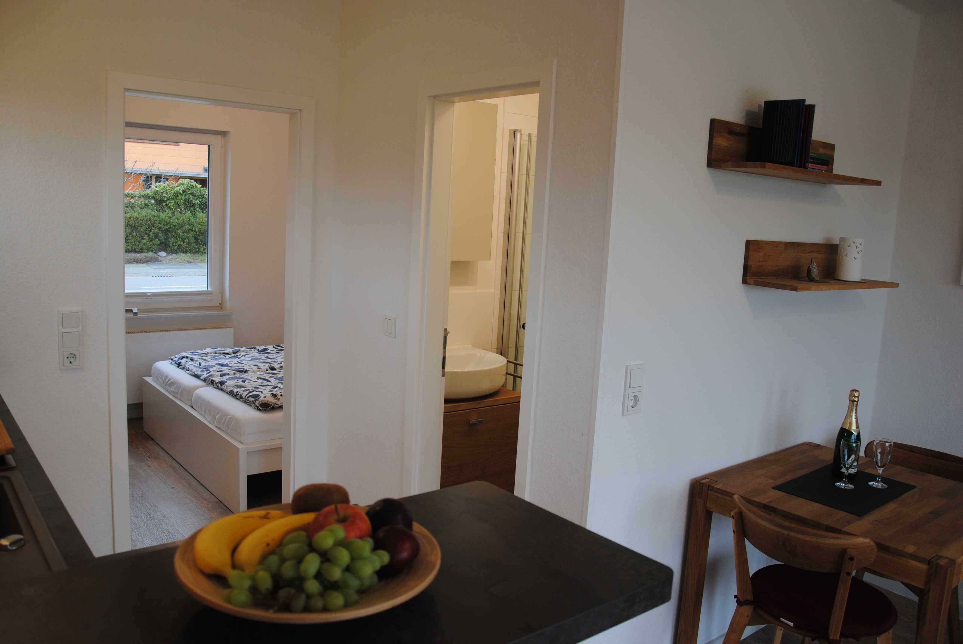 A compact room with a bed, a small table with a fruit bowl, a wooden chair, and a bottle of champagne on a small table.