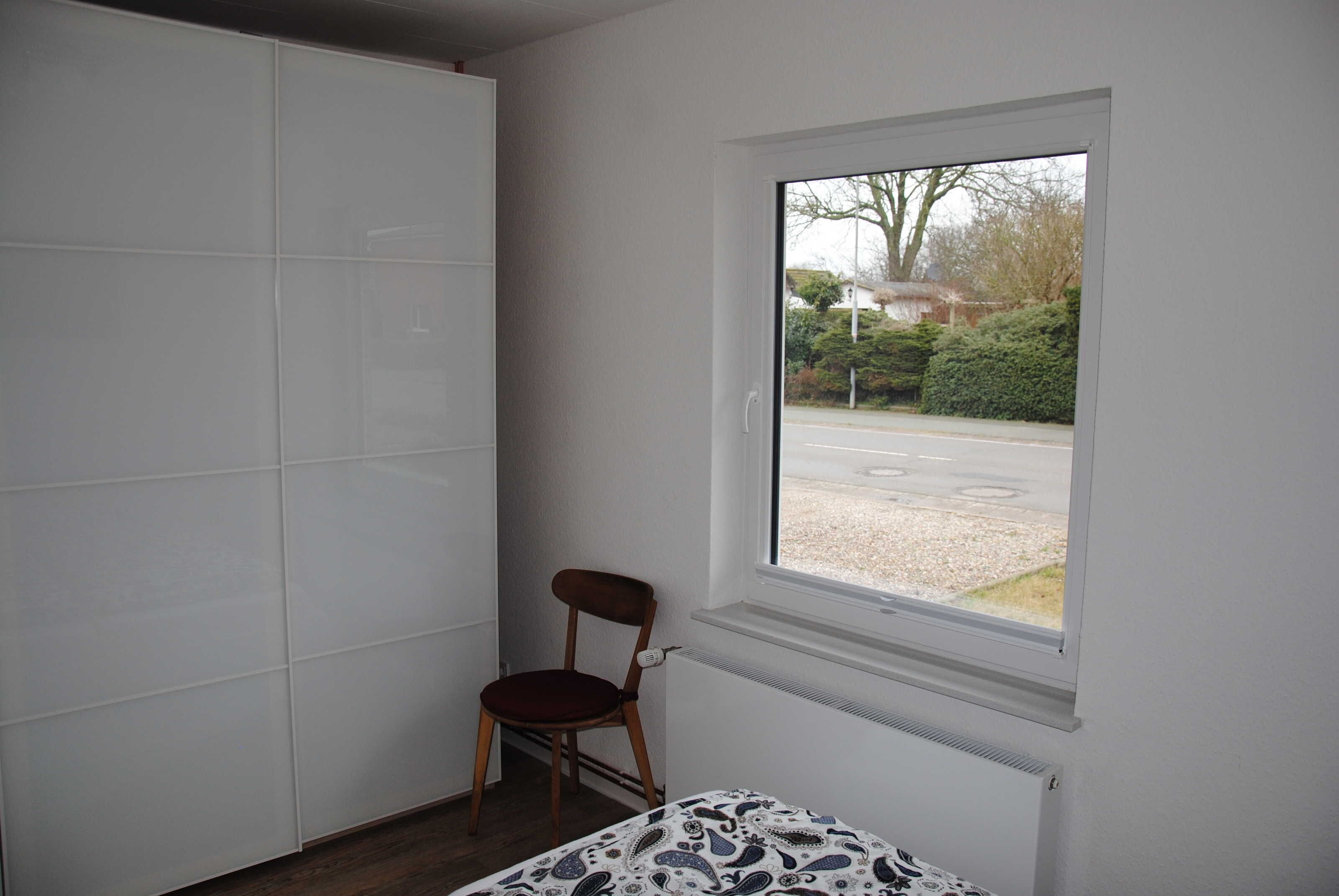 A room with a large window, a radiator, a chair, and a wardrobe.