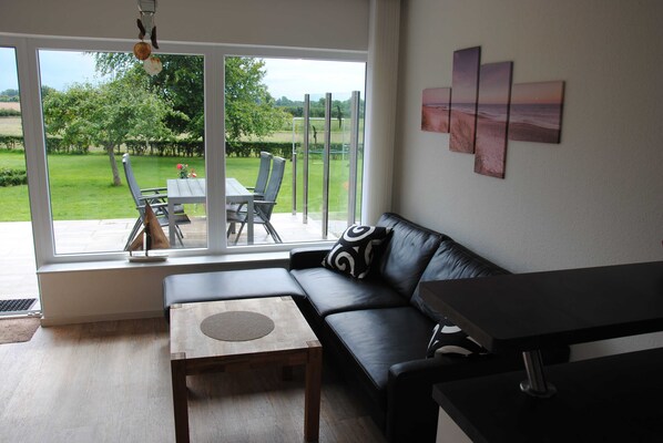 Una sala de estar moderna con un sofá de cuero negro, una mesa de centro de madera y una mesa de comedor con sillas. Amplias ventanas ofrecen vista a un jardín verde y a una playa.
