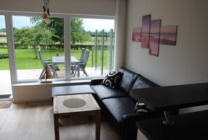 A modern living room with a black leather sofa, a wooden coffee table, and a dining table with chairs. Large windows offer a view of a green garden and a beach.