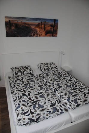 Una cama individual con edredón estampado, cabecera blanca y un cuadro colgado en la pared con una playa.