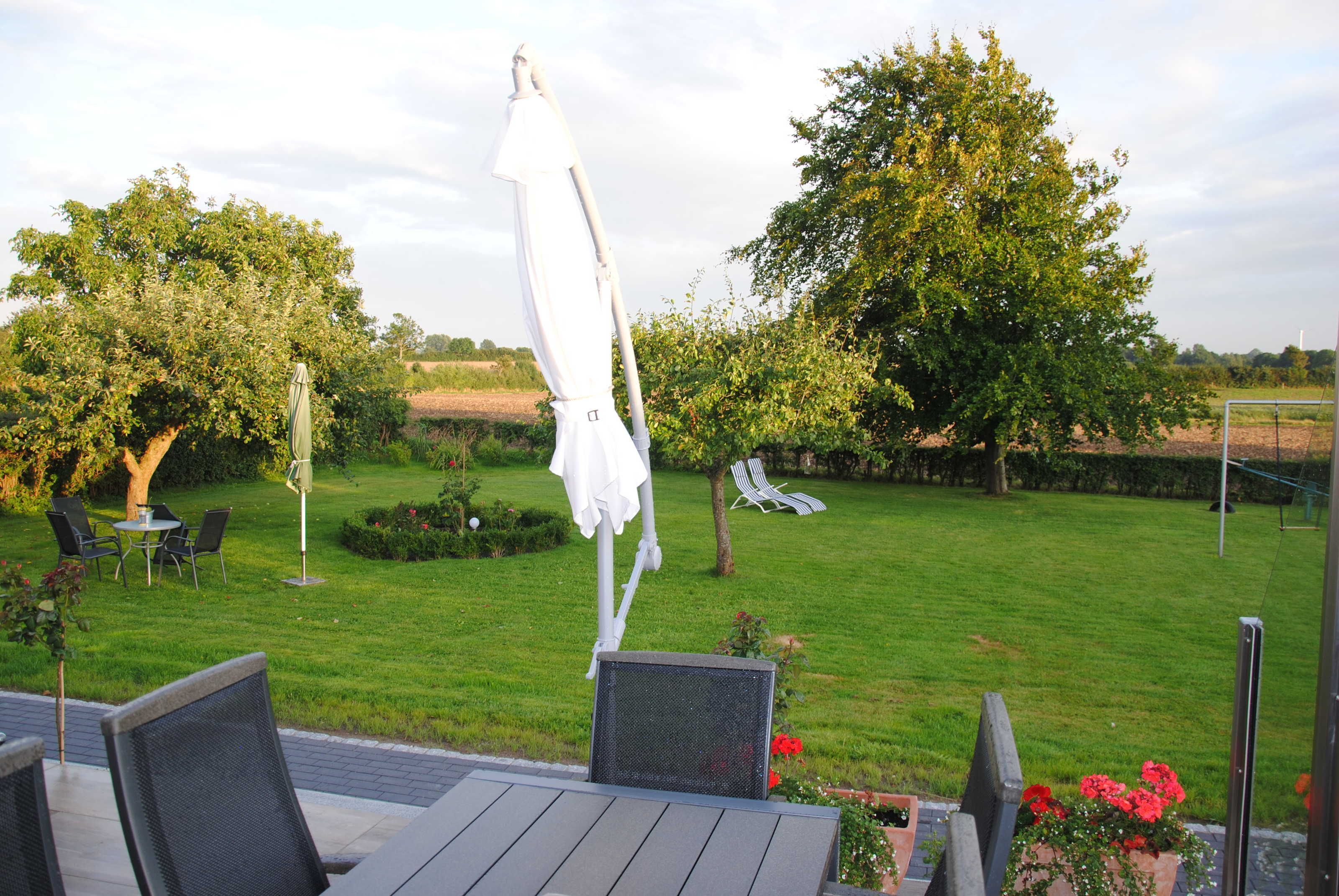 An outdoor patio with a table and chairs, a white umbrella, and a view of a grassy area with trees and a fence.