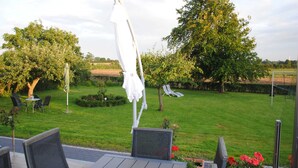 An outdoor patio with a table and chairs, a white umbrella, and a view of a grassy area with trees and a fence. - Vacation apartment in Wackerballig/Gelting 200m to the beach - Holiday apartment (Gelting)