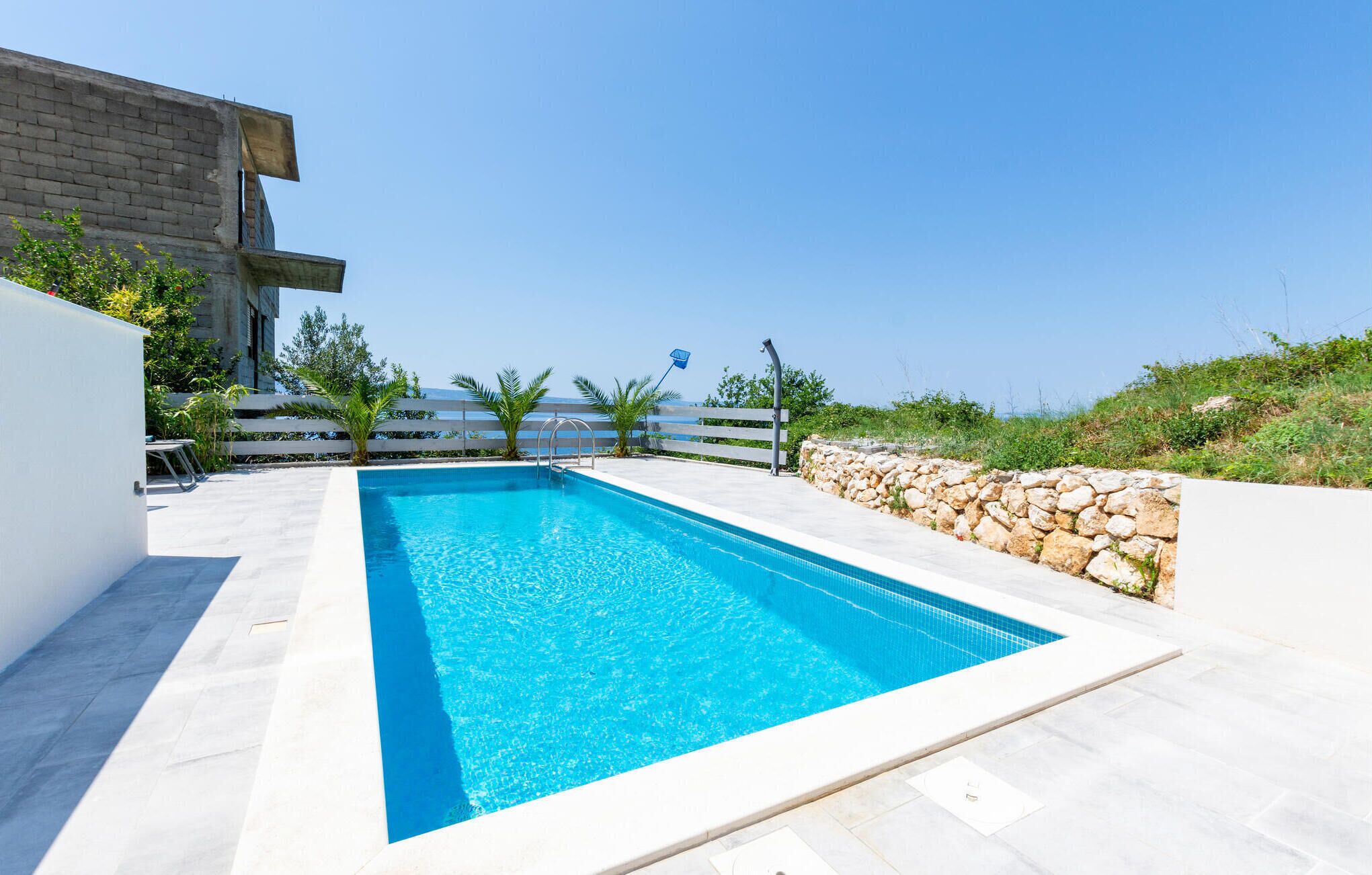 A rectangular swimming pool with clear blue water, surrounded by a white tiled deck and a stone wall.