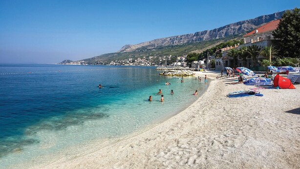 Une plage à l’eau turquoise et limpide, un rivage sablonneux et, en arrière-plan, une colline rocheuse.