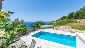 Une piscine avec vue sur la mer, entourée de verdure et bordée d’un mur de pierre.