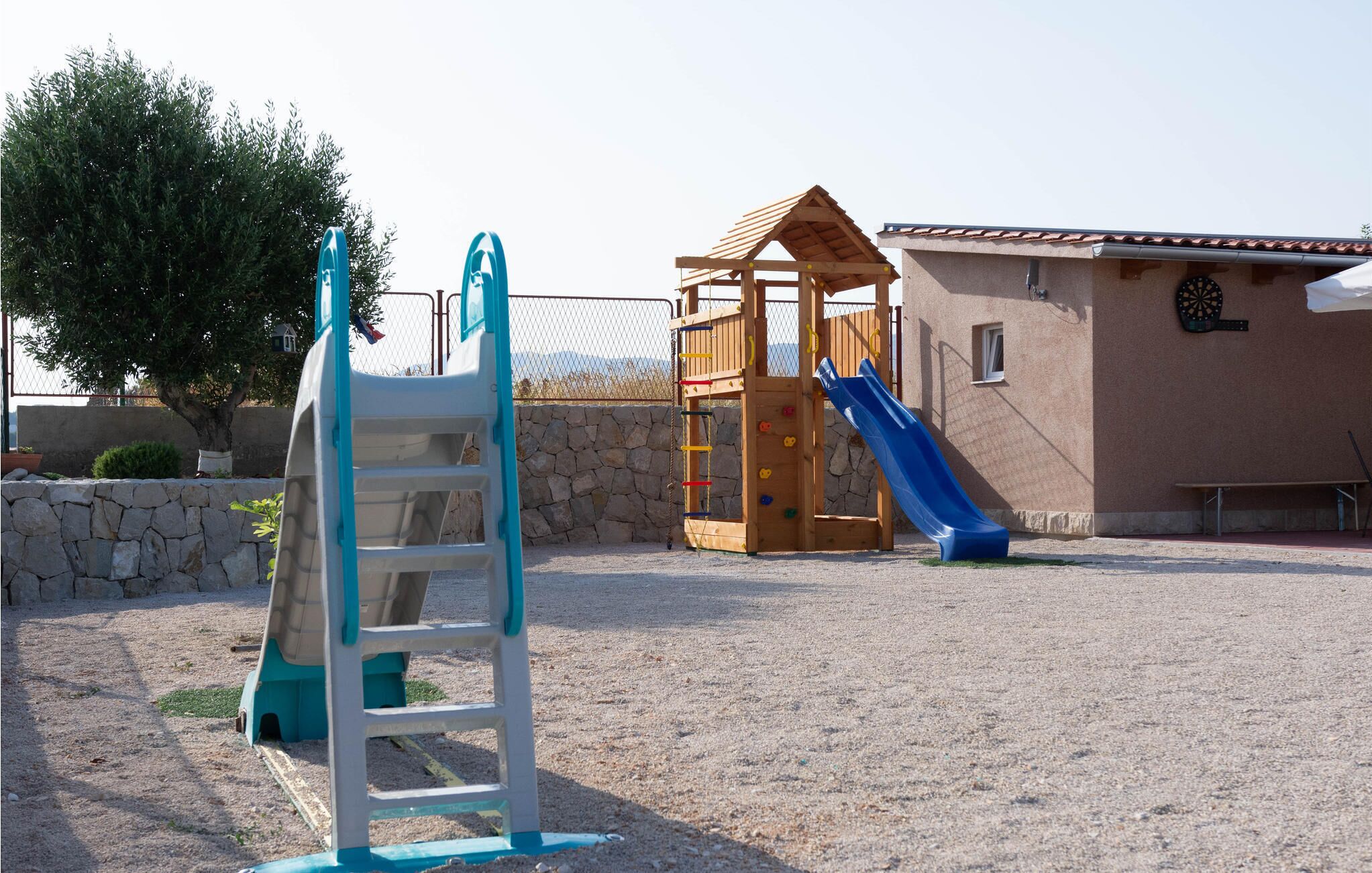 Un terrain de jeux équipé d’un toboggan et de structures d’escalade, entouré d’un mur en pierre et bordé d’un bâtiment dont le toit est recouvert de tuiles.