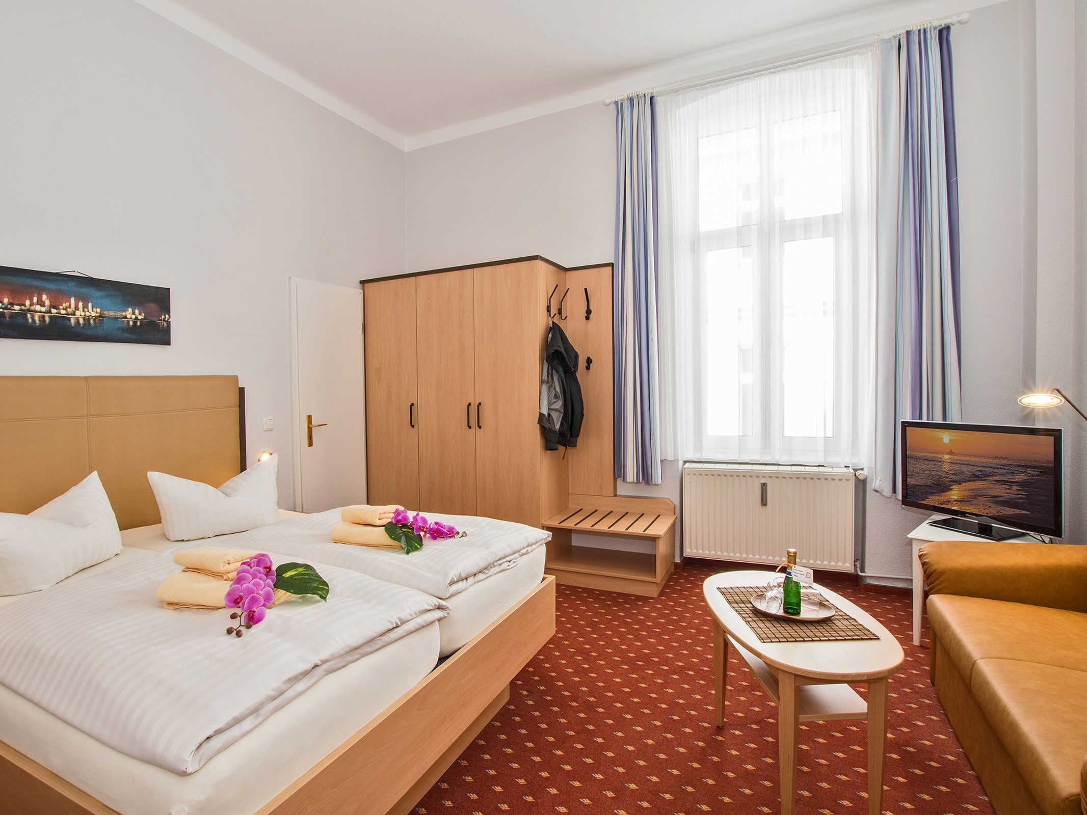 Ein Hotelzimmer mit zwei Betten, einem Holzschrank, einem kleinen Tisch mit einer Flasche, einem Fernseher und einem Fenster mit Vorhängen.