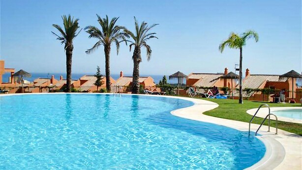A large swimming pool with a curved edge, surrounded by palm trees and beach houses with thatched roofs. - Awesome apartment in Elviria with WiFi (Elviria)