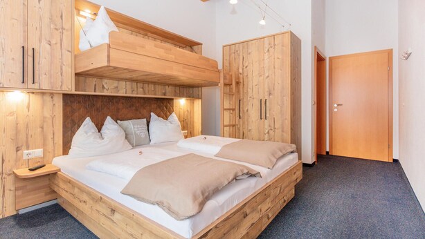 A wooden bunk bed room with a loft bed and a single bed, both with white bedding. There are wooden wardrobes and a door visible.