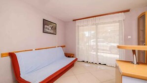 Una habitación pequeña con una cama individual, una ventana con cortinas traslúcidas, una mesita y un espejo.