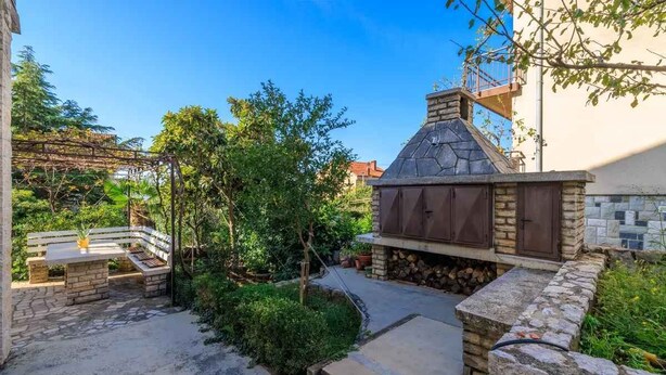 Un área de barbacoa de piedra con chimenea, rodeada de vegetación y una pérgola.