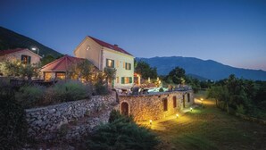 Une maison en pierre avec un toit rouge et un espace piscine, entourée de montagnes et éclairée par un éclairage d’ambiance.