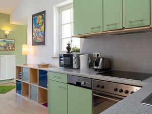 A kitchen with green cabinets, a stove, a kettle, and a toaster. There is a window with a view of the outside, and a painting on the wall.