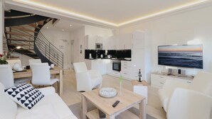 A modern living room with a staircase, a kitchen with white cabinets, and a large TV displaying a sunset.