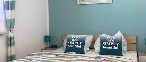 Une chambre à coucher avec un lit, deux oreillers, une table de chevet, une lampe, un mur orné d’un tableau et une fenêtre avec des rideaux.