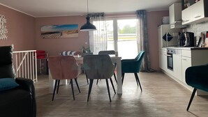 Ett modernt kök med vita skåp, en matplats med bord och stolar, ett stort fönster med gardiner och en takfläkt.