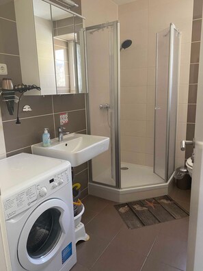 Un baño con lavadora, lavamanos, cabina de ducha de vidrio y paredes de azulejos.